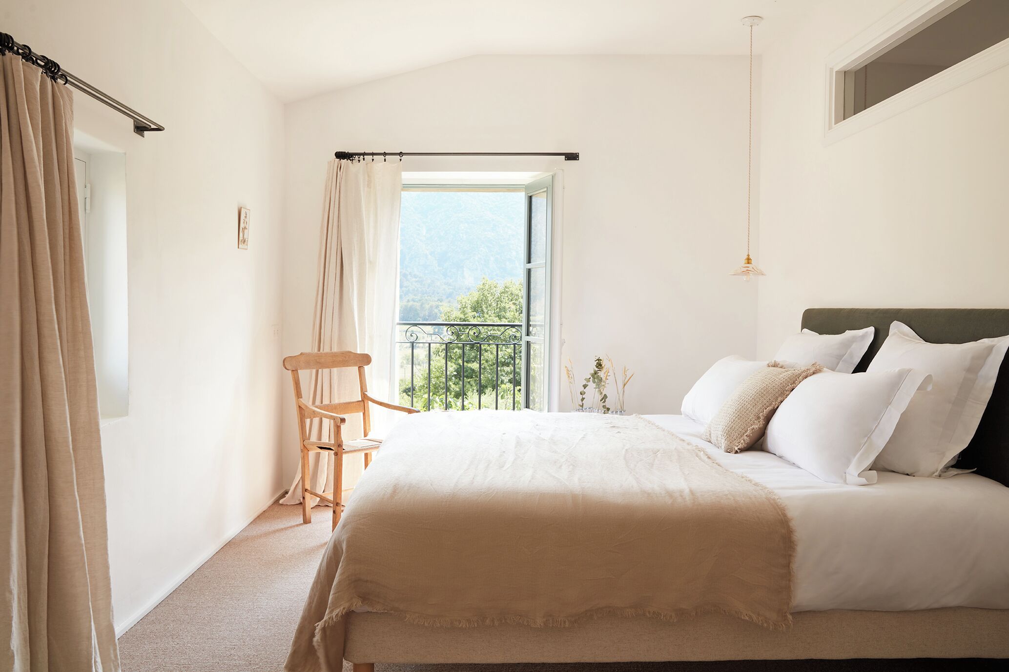 A bedroom with a bed, a chair, a balcony with a view, and a hanging light fixture.