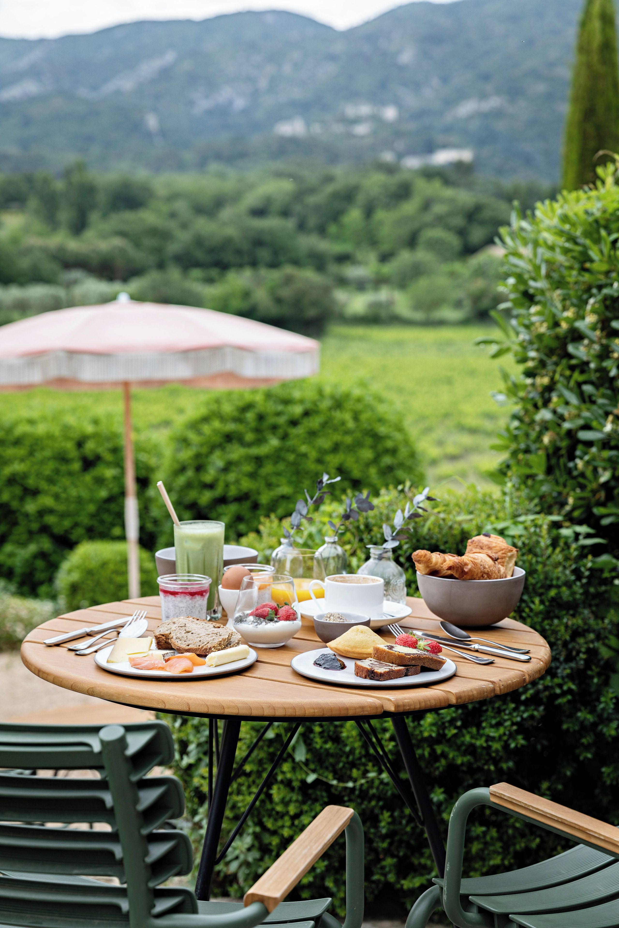 An outdoor dining table with a variety of food and drinks, surrounded by greenery and a mountain view.