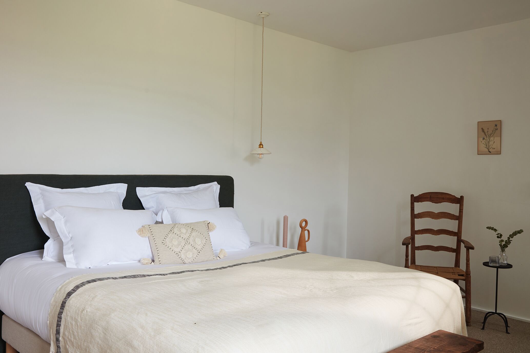A neatly made bed with white bedding, a wooden chair, and a small table with a vase.