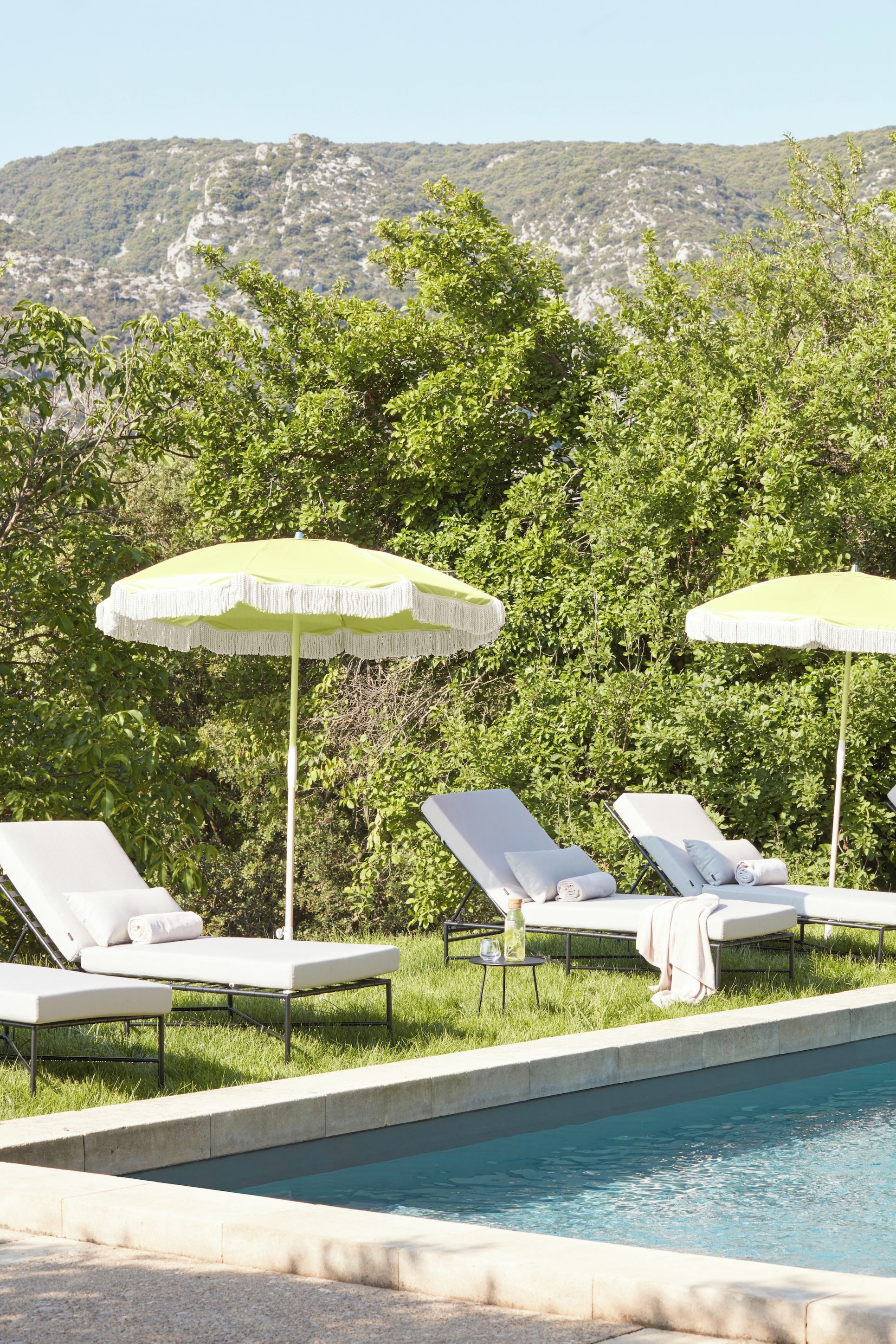 A poolside area with lounge chairs and umbrellas, surrounded by greenery and a mountainous backdrop.