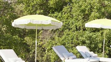 A poolside area with lounge chairs and umbrellas, surrounded by greenery and a mountainous backdrop.