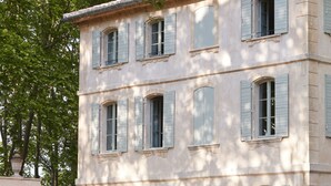 A large, light-colored building with multiple windows and shutters, surrounded by a well-maintained garden with hedges.