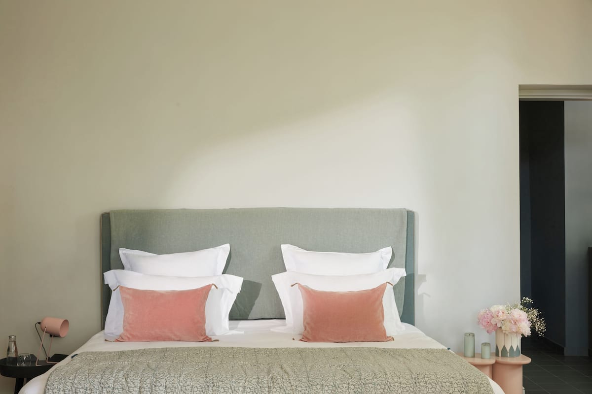 A neatly made bed with a grey headboard, white and pink pillows, and a patterned bedspread. There is a small bedside table with a lamp and a vase of flowers.
