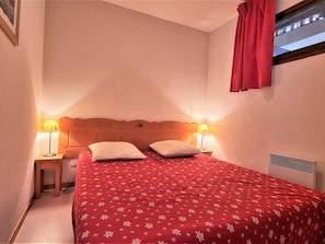 Quarto com cabeceira de madeira, duas luminárias de cabeceira, colcha vermelha com estampa floral e janela com cortina vermelha.