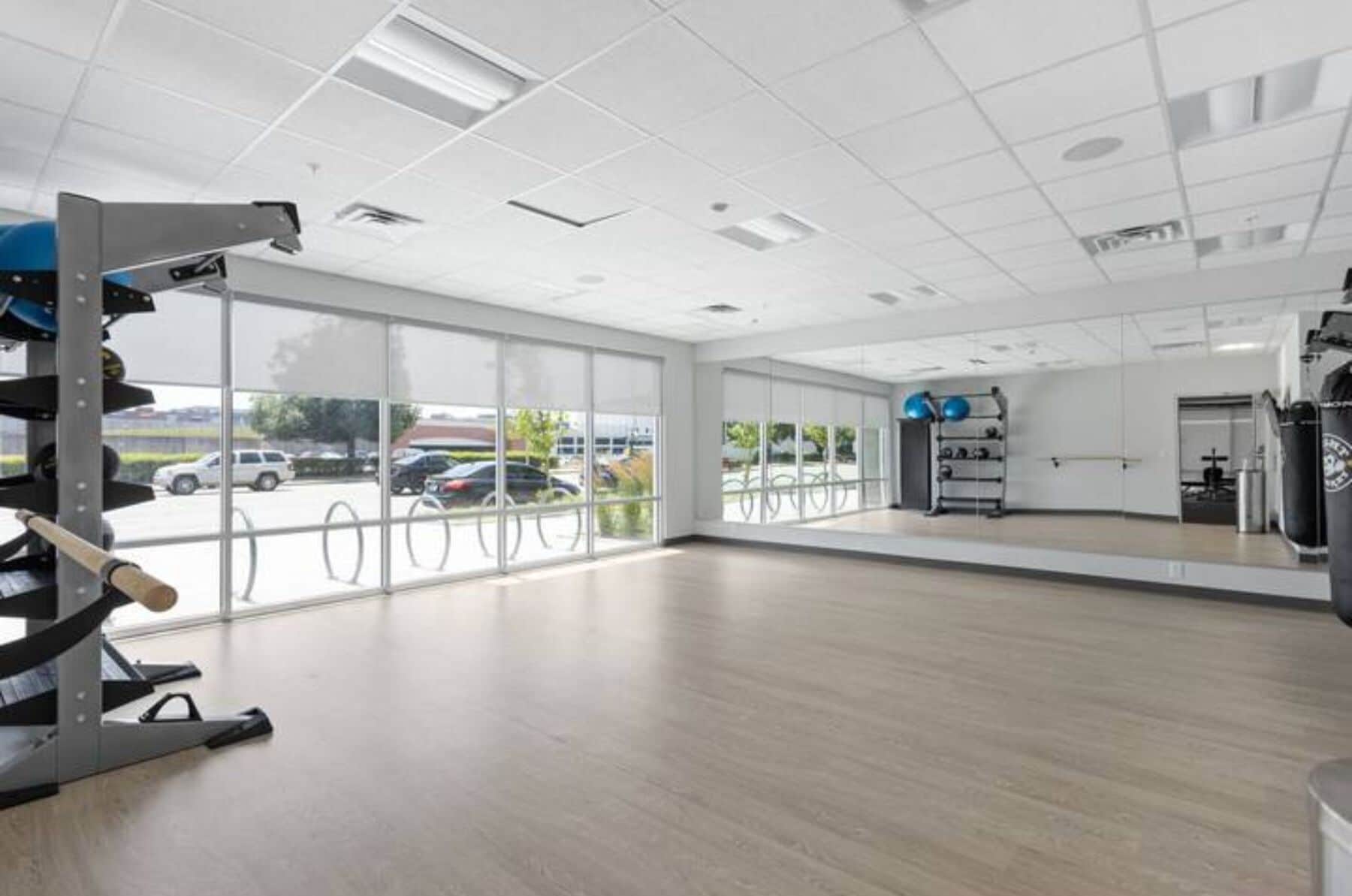 A spacious gym with large windows, exercise equipment, and a view of parked cars outside.