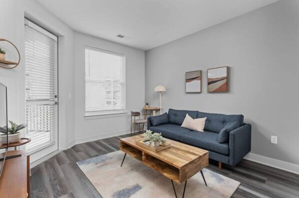 A modern living room with a blue sofa, wooden coffee table, and a large window with blinds.
