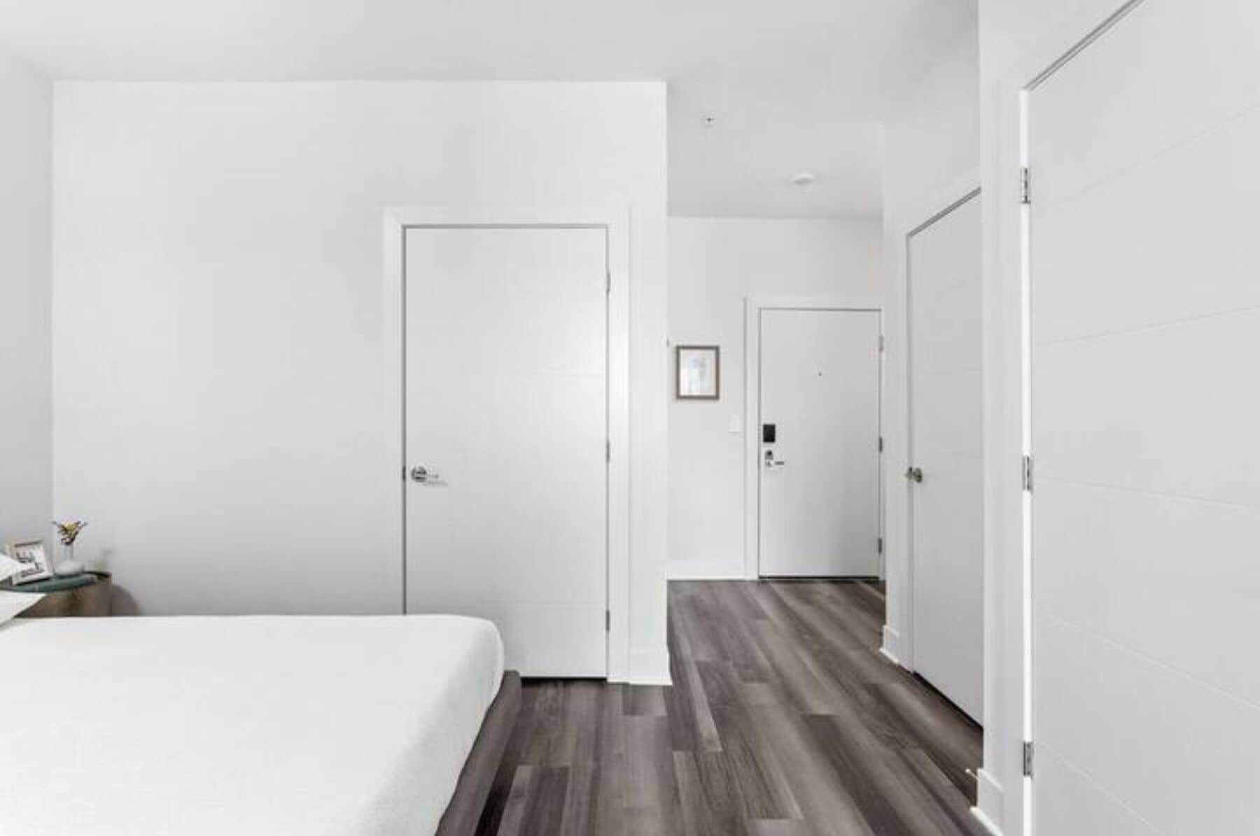 Habitación de hotel con una cama grande, suelo de madera y paredes blancas. Se ve una puerta que da a otra habitación y una mesita con una lámpara.