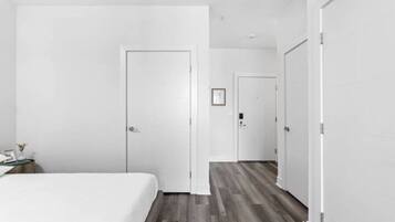 Habitación de hotel con una cama grande, suelo de madera y paredes blancas. Se ve una puerta que da a otra habitación y una mesita con una lámpara.