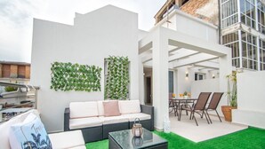 A modern outdoor living area with a white wall, green plants, a white sofa, a glass coffee table, and a dining set. - Alcmene Lux Apartment (Zakynthos)