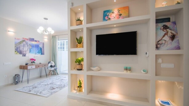 A modern living room with a flat-screen TV mounted on a white wall, surrounded by shelves with decorative items and plants. - Alcmene Lux Apartment (Zakynthos)