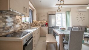 A modern kitchen with a built-in stove, a dining area with a table and chairs, and a living area with a sofa and a clock on the wall. - Alcmene Lux Apartment (Zakynthos)