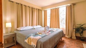 A hotel room with a bed, curtains, a nightstand, a lamp, and a potted plant.