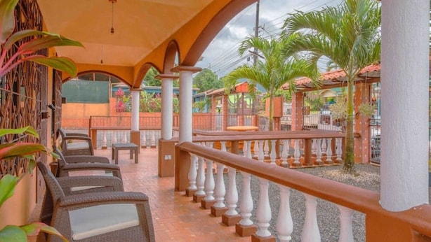 A covered outdoor seating area with wicker furniture and a railing, surrounded by palm trees and a brick wall.