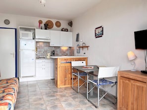 Uma cozinha compacta com micro-ondas, geladeira e armários de madeira. Há um pequeno espaço de jantar com mesa e cadeiras.