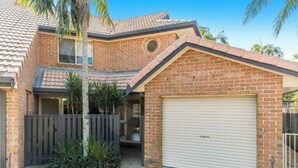 A two-story brick house with a garage, a fenced yard, and a small garden area. - Bayside Court - Three Bedroom (Byron Bay)