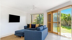A modern living room with a blue sofa, a flat-screen TV, and a sliding glass door leading to a garden. - Bayside Court - Three Bedroom (Byron Bay)