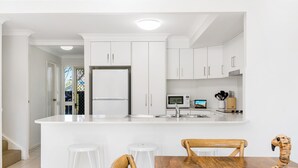 A modern kitchen with white cabinetry, a central island with stools, and a wooden dining table. - Bayside Court - Three Bedroom (Byron Bay)
