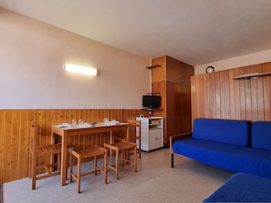 Quarto compacto com mesa de madeira posta para refeições, um sofá azul e uma pequena televisão num armário.