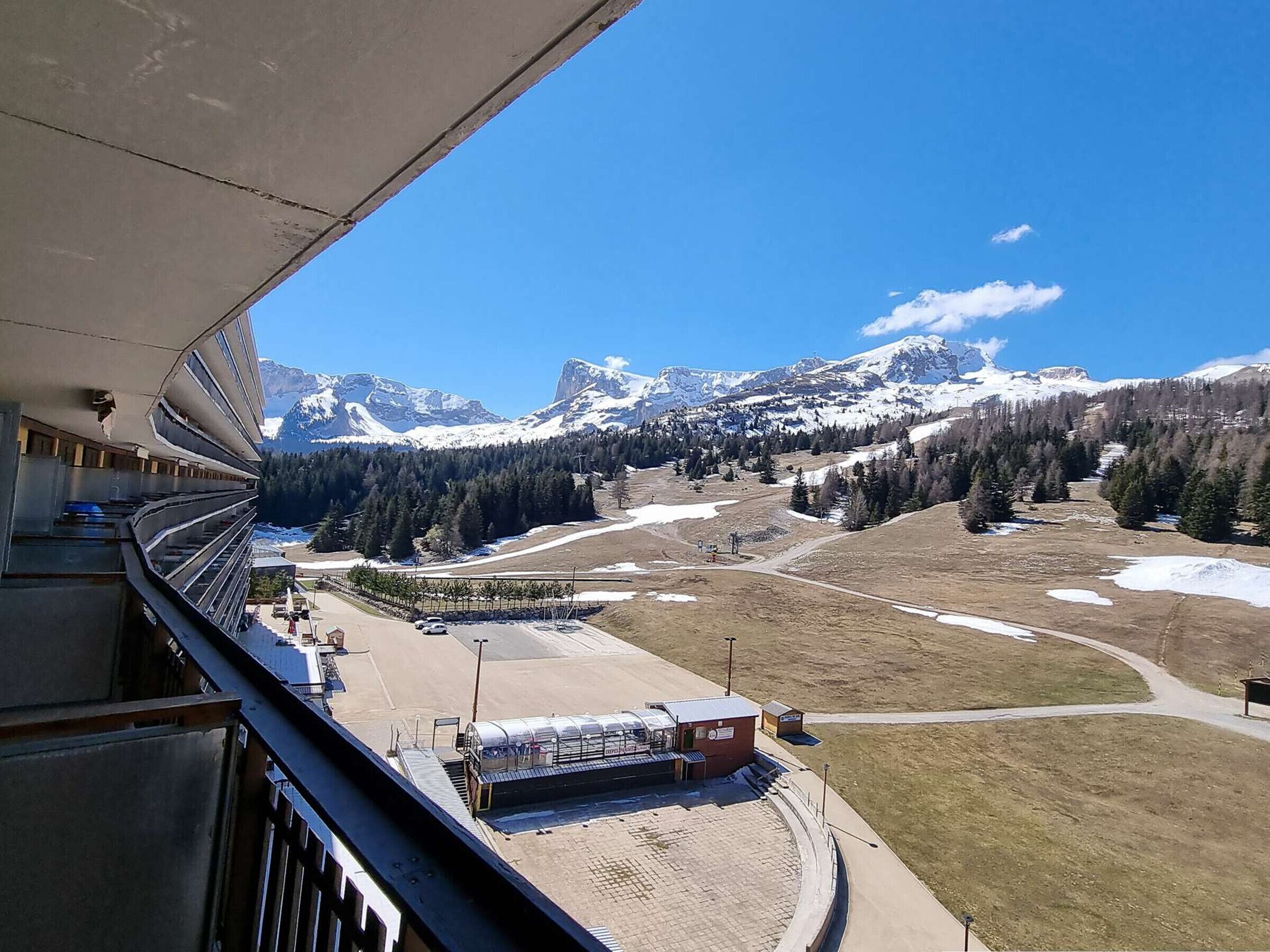 A view from a balcony overlooking a mountainous landscape with snow-capped peaks, a ski resort, and a clear blue sky.