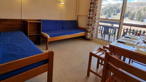 A room with a bed, a sofa, a dining table with chairs, and a view of a snowy mountain landscape through a large window. - Cozy studio for 4 with terrace, near slopes and shops (Le Dévoluy)