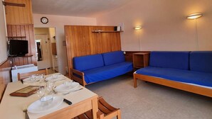 A room with a dining table set for four, a blue sofa, a wooden cabinet, and a television mounted on the wall. - Cozy studio for 4 with terrace, near slopes and shops (Le Dévoluy)