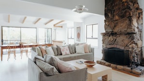 A spacious living room with a stone fireplace, a large window, and a dining area with wooden chairs. - Massinger Views Byron Bay (Byron Bay)