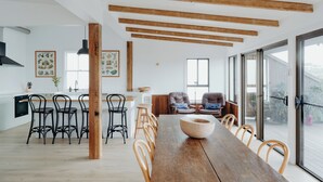 A spacious dining area with a long wooden table and chairs, a kitchen with white cabinets and black chairs, and large windows offering views of the outdoors. - Massinger Views Byron Bay (Byron Bay)