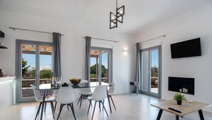 Un espace repas moderne avec une table en verre, des chaises blanches et une télécommande posée sur une petite table d’appoint en bois.