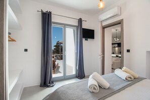 Ein Schlafzimmer mit Bett, einem Fenster mit Vorhängen, einem an der Wand befestigten Fernseher und einem durch die Tür einsehbaren Badezimmer.