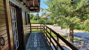 A wooden balcony with a metal railing, a door with glass panels, and a view of a road and greenery.
