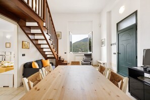 A dining area with a wooden table, chairs, a sofa with cushions, a staircase, and a view through a window.