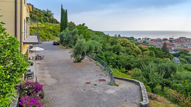 Un sentier pavé de pierres mène à un bâtiment doté d’une terrasse, entouré de verdure et offrant une vue sur une ville côtière.