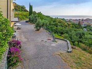 A cobblestone pathway leading to a building with outdoor seating, surrounded by greenery and overlooking a coastal town.