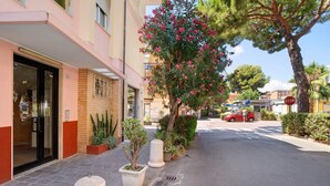 A building with a glass entrance, potted plants, a tree with pink flowers, and a red car parked on the street. - Apartment 'Mari Fior' with Sea View, Balcony and Wi-Fi (San Bartolomeo)