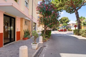 A building with a glass entrance, potted plants, a tree with pink flowers, and a red car parked on the street.