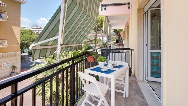 A balcony with a table and chairs, a potted plant, and a view of the street and buildings. - Apartment 'Mari Fior' with Sea View, Balcony and Wi-Fi (San Bartolomeo)