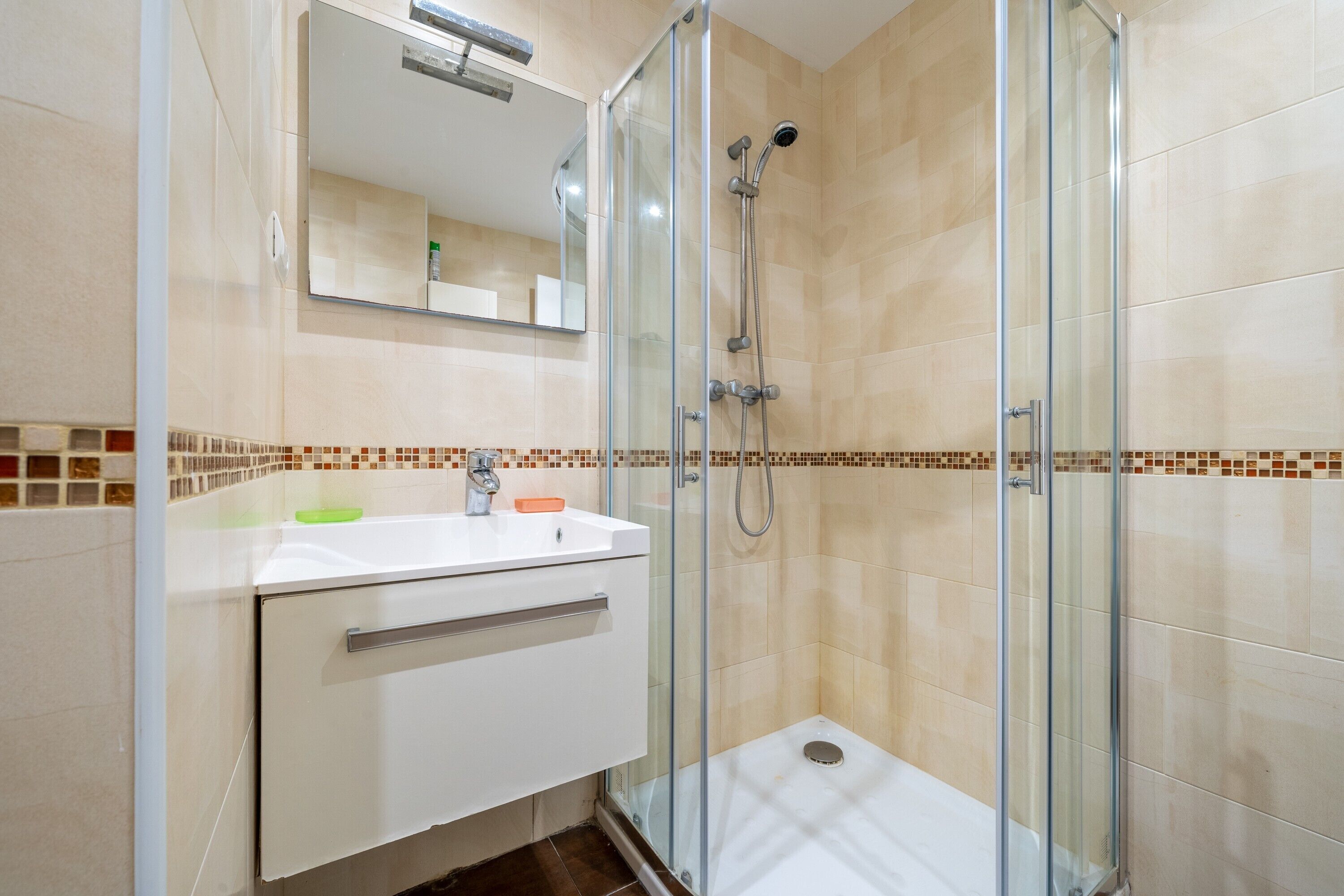 Uma casa de banho moderna com cabine de duche em vidro, lavatório branco com espelho em cima e chão em mosaico.
