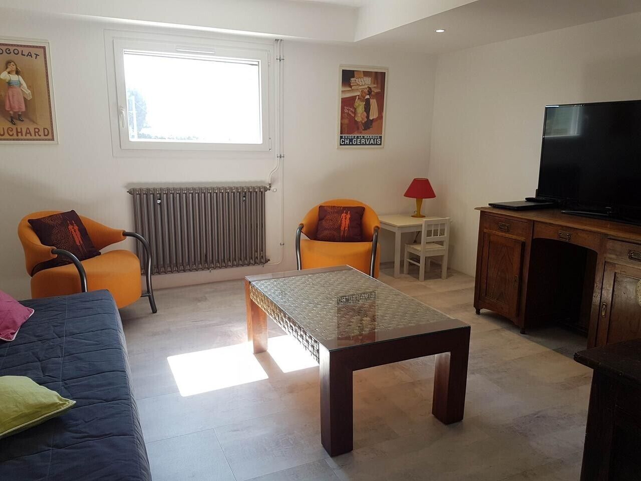 A living room with a sofa, two armchairs, a glass-top coffee table, a TV, and a radiator.