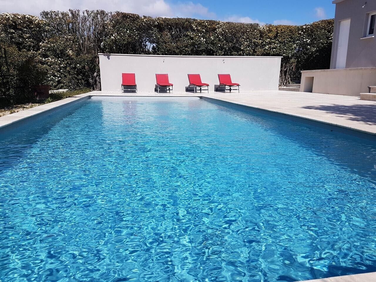 A rectangular swimming pool with clear blue water, surrounded by a white wall and a row of red lounge chairs.
