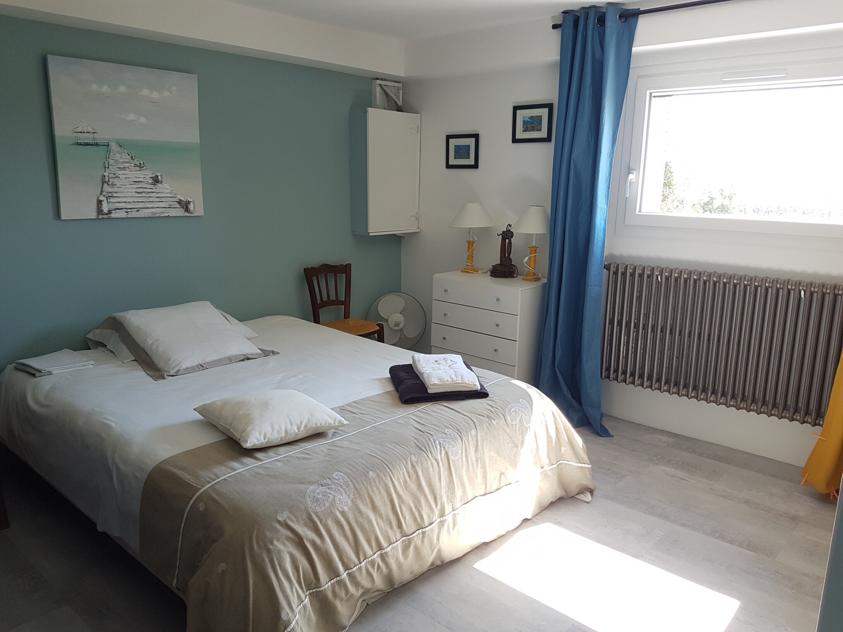 Une chambre à coucher comprenant un lit, une chaise, une commode, un ventilateur, un radiateur et une fenêtre avec des rideaux.