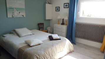 Une chambre à coucher comprenant un lit, une chaise, une commode, un ventilateur, un radiateur et une fenêtre avec des rideaux.
