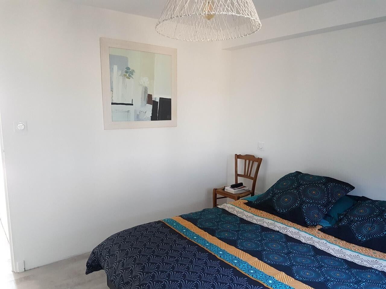 Un dormitorio con cama, una silla, una obra de arte enmarcada en la pared y una lámpara colgante.