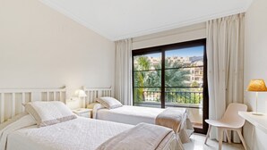 A hotel room with two beds, a desk, a chair, a window with a view of palm trees and buildings, and a lamp. - Apartment 'Villa Gadea 4-3' with Sea View, Wi-Fi and Air Conditioning (ALTEA)