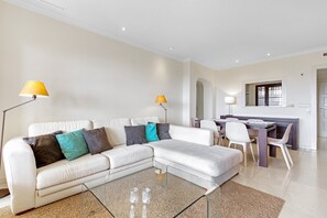 Una sala de estar moderna con un sofá seccional blanco, dos lámparas de pie, una mesa de centro de vidrio y una zona de comedor con una mesa de madera oscura y sillas blancas.
