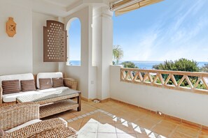 Ein Balkon mit Meerblick, eine Rattan-Couch, ein Glastisch und ein dekorativer Sichtschutz.