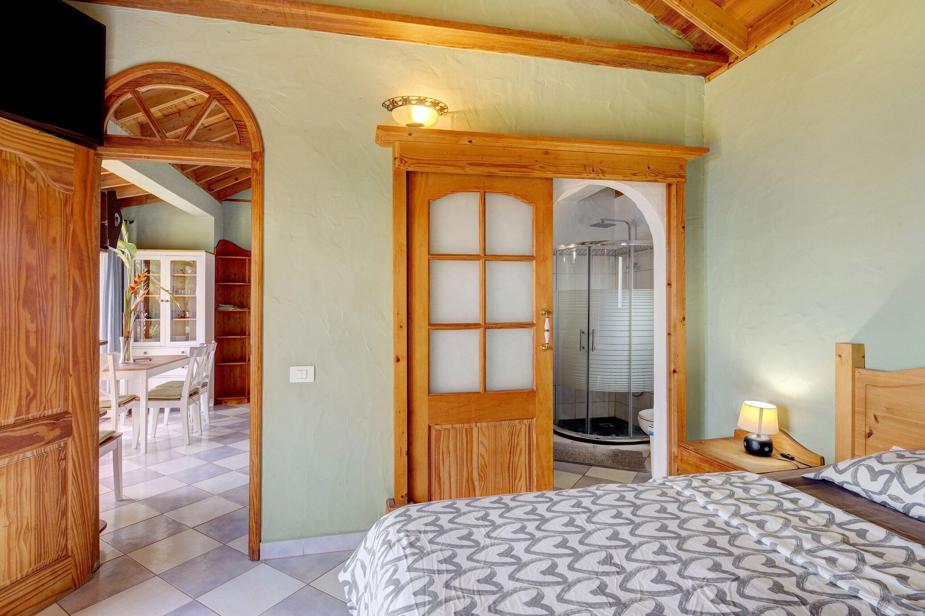 A bedroom with a bed, a wooden door, a shower, and a dining area with chairs and a table.