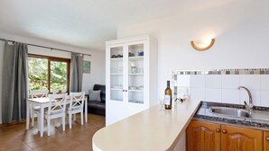Una cocina con encimera blanca, un lavabo y armarios. Hay una mesa de comedor con sillas y una ventana con cortinas.