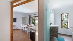 Una habitación con viga de madera en el techo, comedor con mesa y sillas, un armario blanco y ventana con vistas a la vegetación.