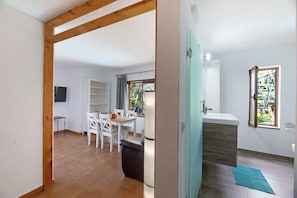 A room with a wooden ceiling beam, a dining area with a table and chairs, a white cabinet, and a window with a view of greenery.