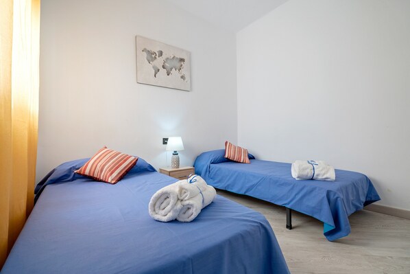 A hotel room with two beds, each with blue bedding and a rolled white towel. There is a world map on the wall, a bedside lamp, and a small table with a lamp.